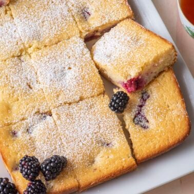 Blackberry tea cake cut into squares and dusted with powdered sugar on a white platter with fresh blackberries and a cup of tea.