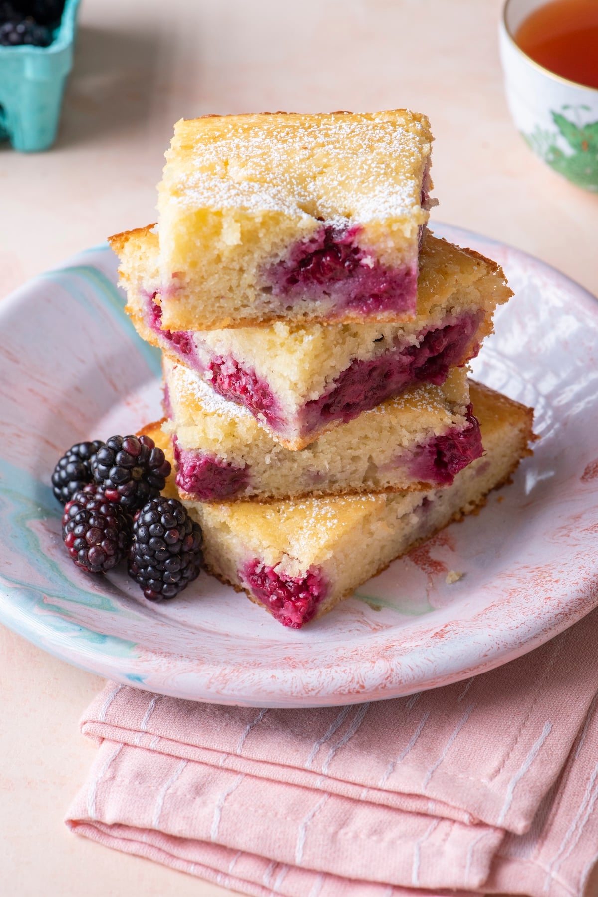 Three squares of blackberry tea cake stacked on a plate showing the blackberry filling inside, dusted with powdered sugar and served with fresh blackberries and tea.