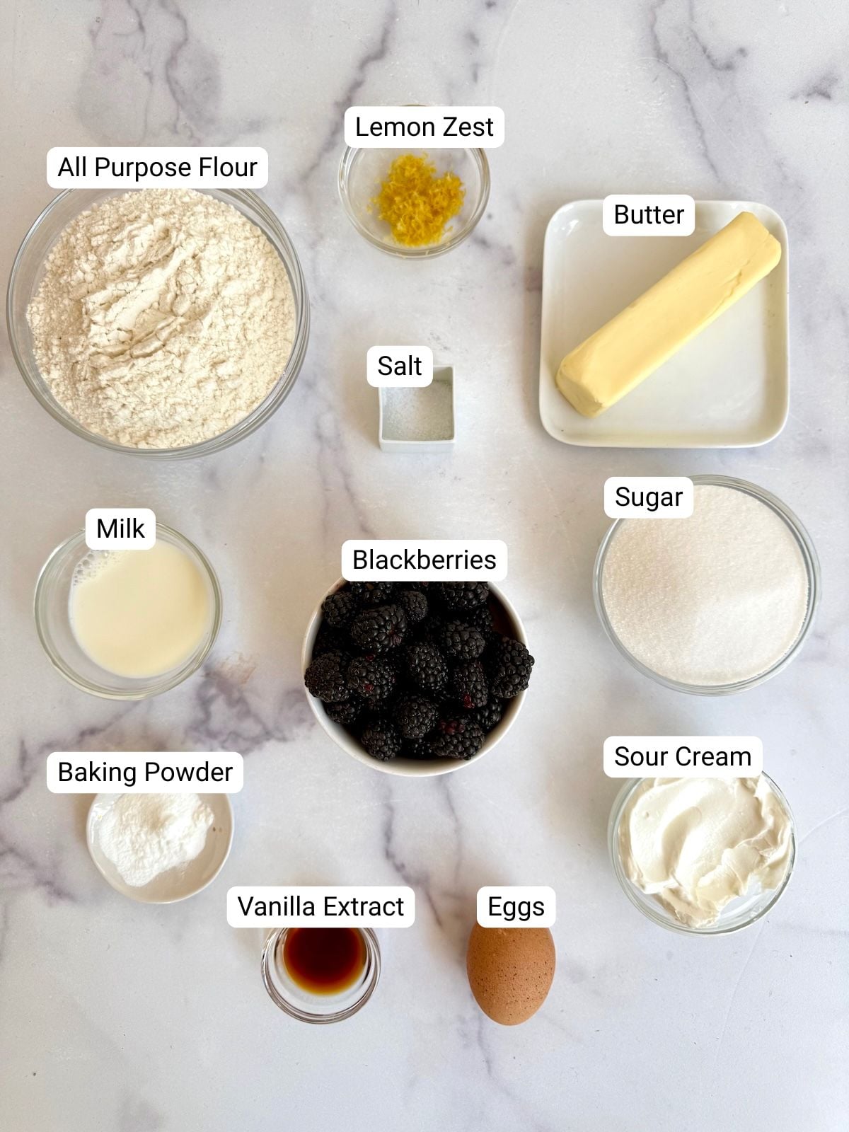 Ingredients for blackberry tea cake including fresh blackberries, flour, butter, sugar, sour cream, eggs, lemon zest, milk, vanilla, and baking powder arranged on a marble surface.