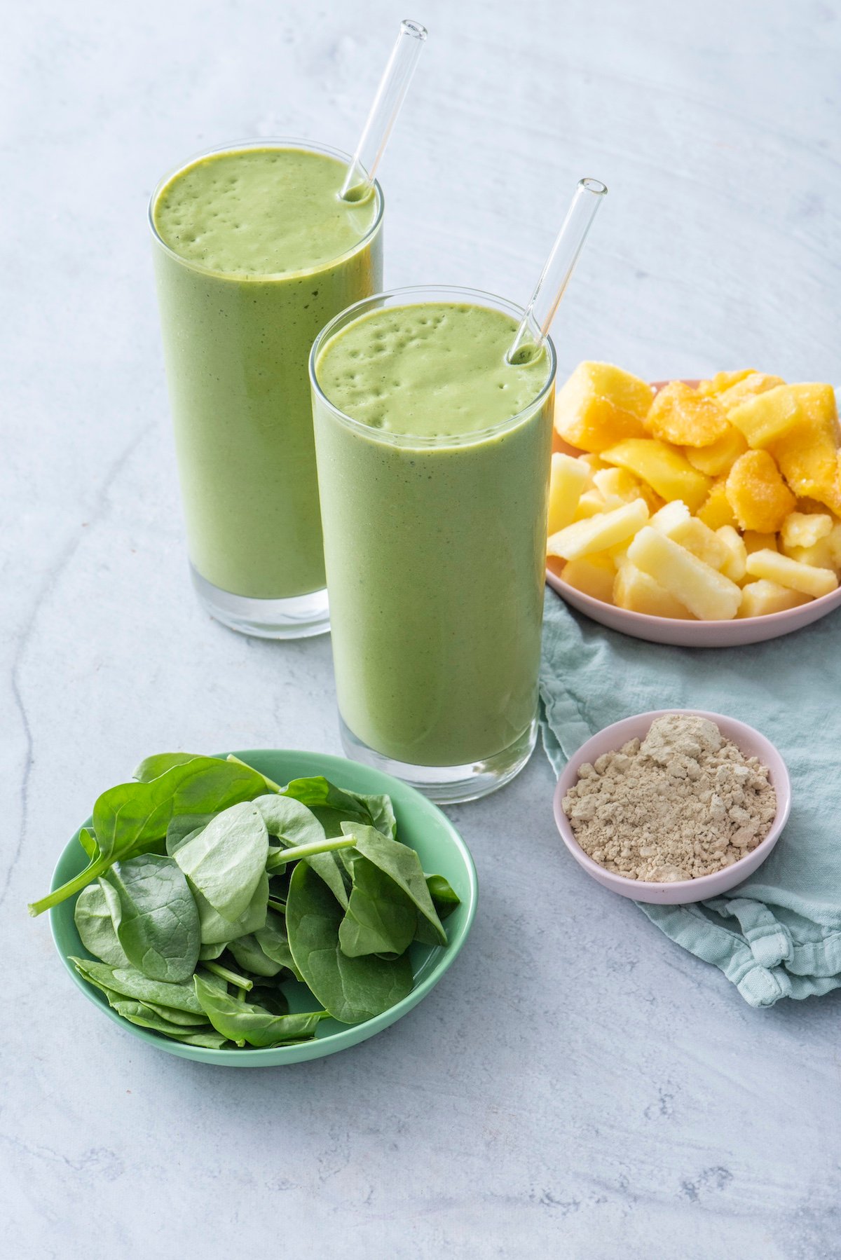 Two glasses filled with green smoothies. Ingredients like mango, pineapple, protein powder and spinach surround the glasses.