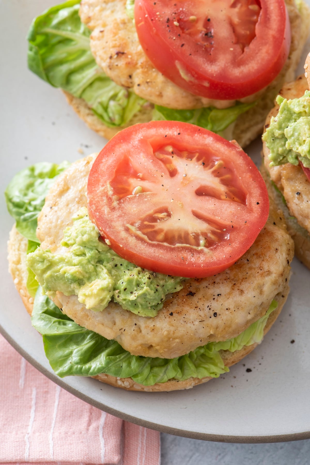 Open-faced juicy turkey burgers on sesame buns topped with guacamole and tomato on a white plate.