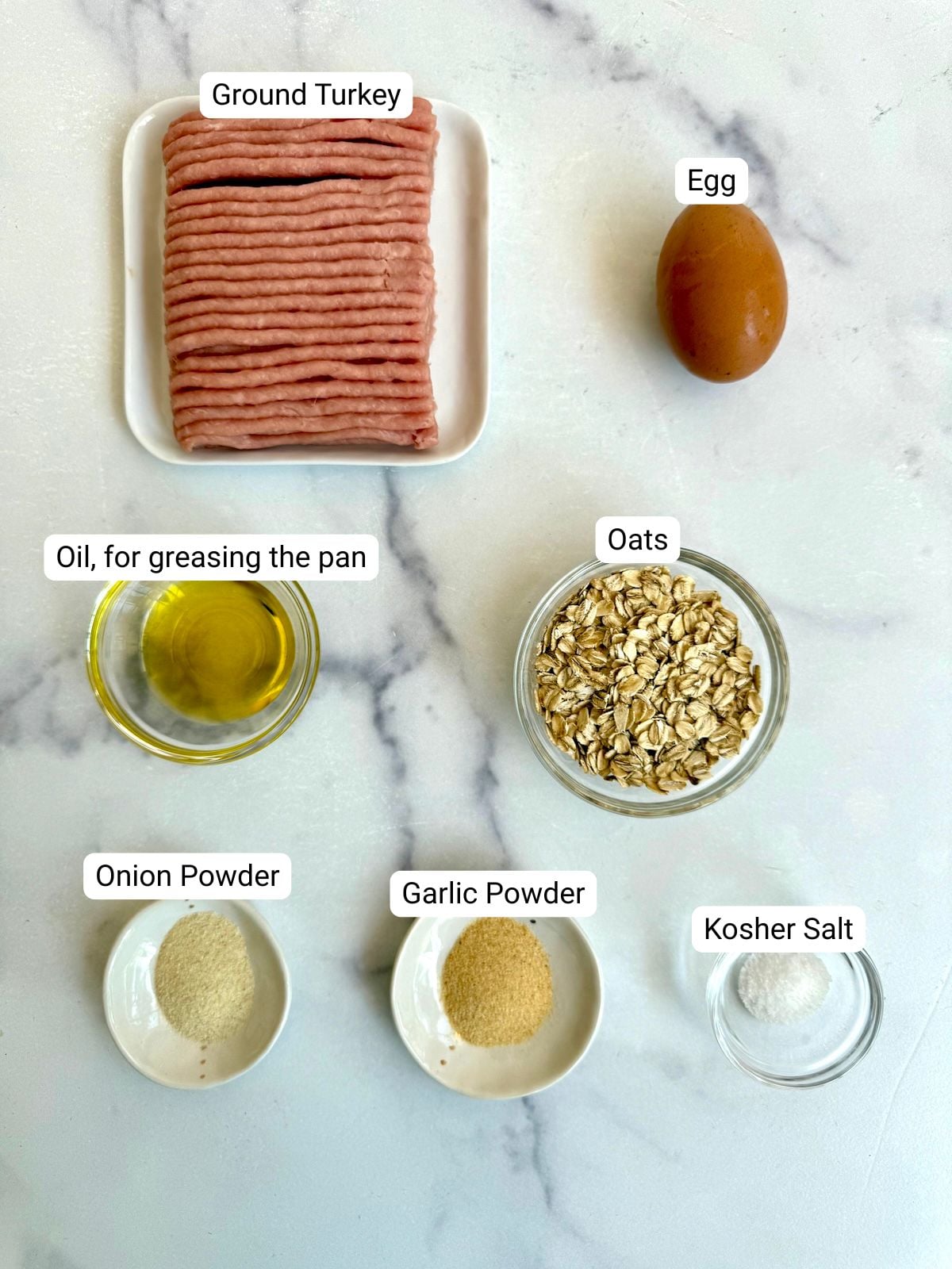Ingredients for juicy turkey burgers laid out on a marble surface: ground turkey, egg, rolled oats, olive oil, garlic powder, onion powder, and kosher salt.