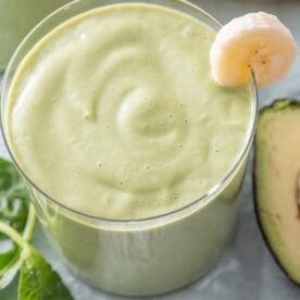 Close-up of a green breakfast smoothie in a glass with a banana slice on the rim, fresh spinach and avocado visible alongside.
