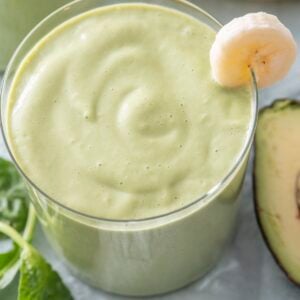 Close-up of a green breakfast smoothie in a glass with a banana slice on the rim, fresh spinach and avocado visible alongside.