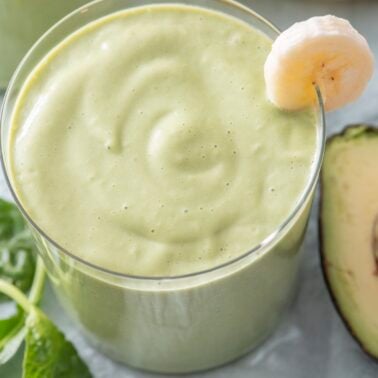 Close-up of a green breakfast smoothie in a glass with a banana slice on the rim, fresh spinach and avocado visible alongside.