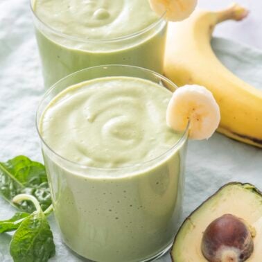 Two glasses of green breakfast smoothie made with avocado, banana, and spinach, garnished with banana slices and served with fresh spinach.