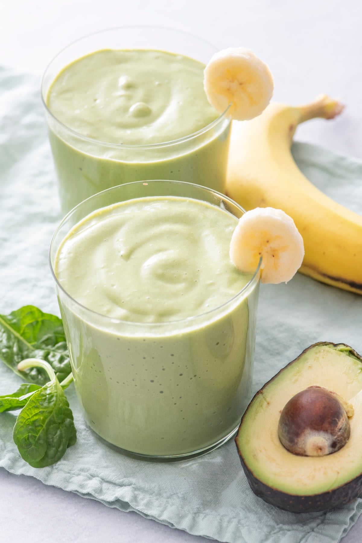 Two glasses of green breakfast smoothie made with avocado, banana, and spinach, garnished with banana slices and served with fresh spinach.