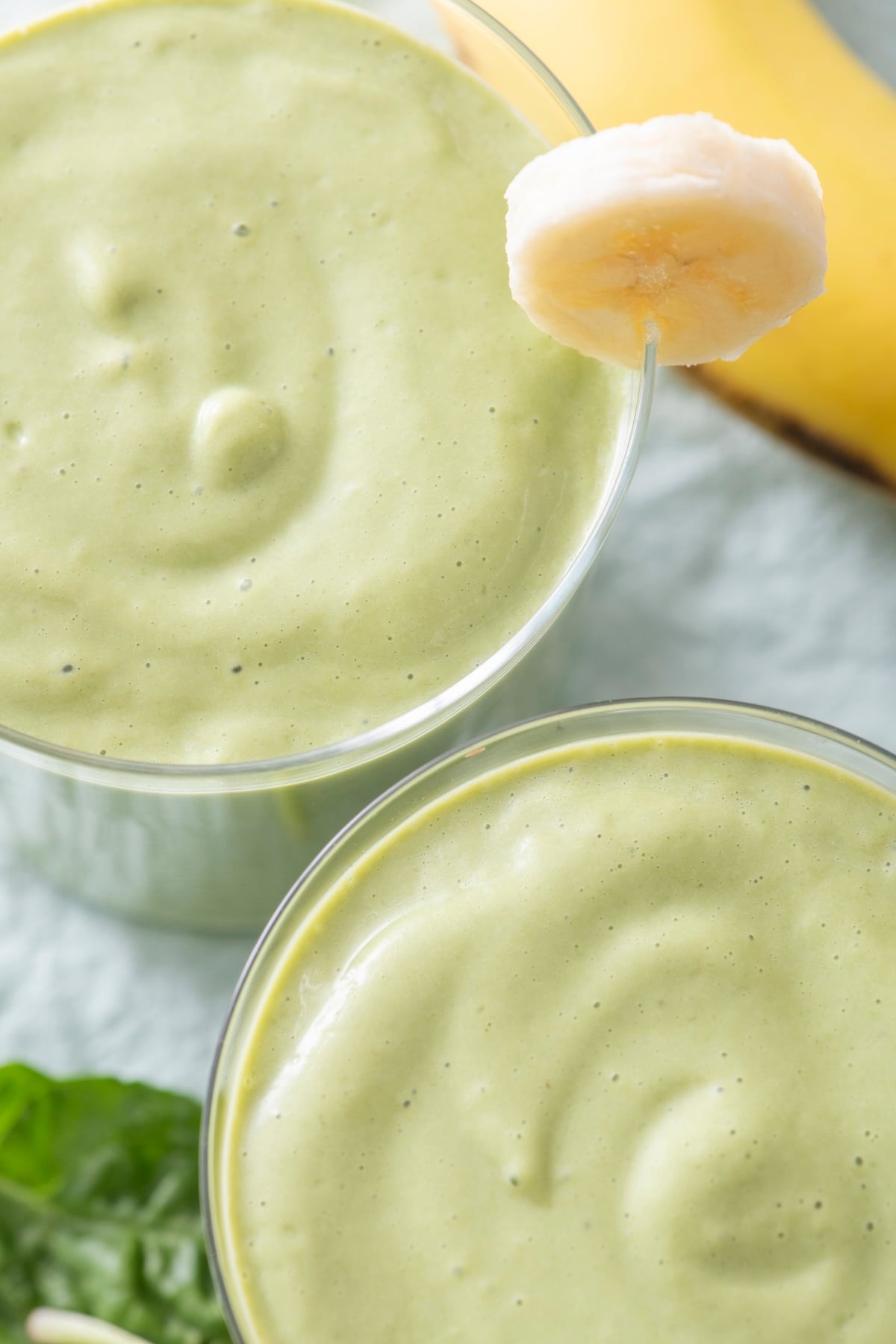 Overhead view of two glasses of creamy green breakfast smoothie with banana slices on the rim and fresh spinach in the background.