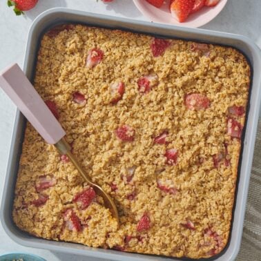A square baking pan with baked strawberry oatmeal, some fresh strawberries, a bowl of sliced strawberries, and a bowl of oats nearby. A gold spoon rests in the oatmeal.
