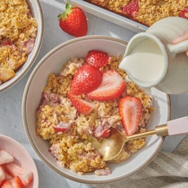 A bowl of baked oatmeal with sliced strawberries on top, a spoon inside, and cream being poured from a small pitcher. A baking dish of more oatmeal and fresh strawberries are visible nearby.
