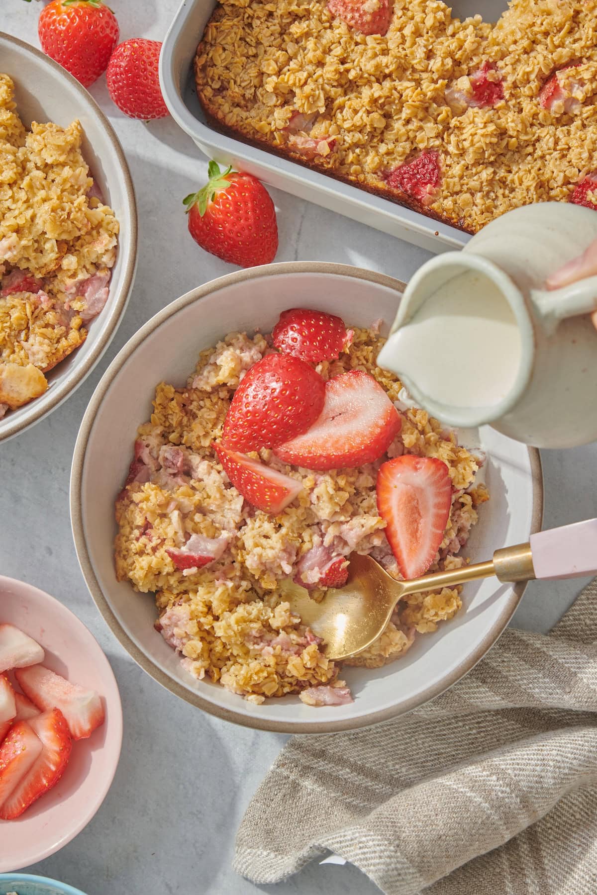 A bowl of baked oatmeal with sliced strawberries on top, a spoon inside, and cream being poured from a small pitcher. A baking dish of more oatmeal and fresh strawberries are visible nearby.