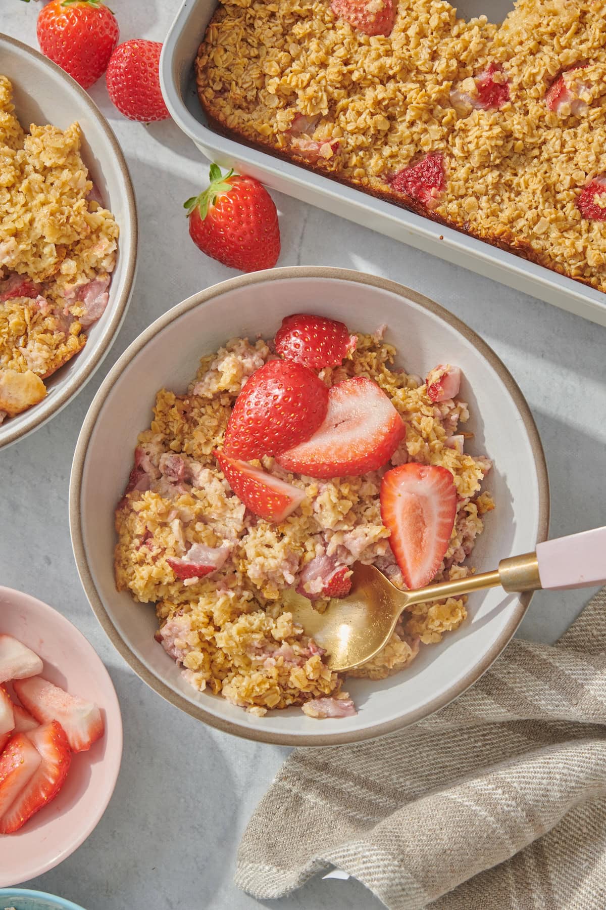 A bowl of baked oatmeal topped with fresh strawberry slices, beside a baking dish of oatmeal and extra strawberries on a light surface. A gold spoon rests in the bowl.
