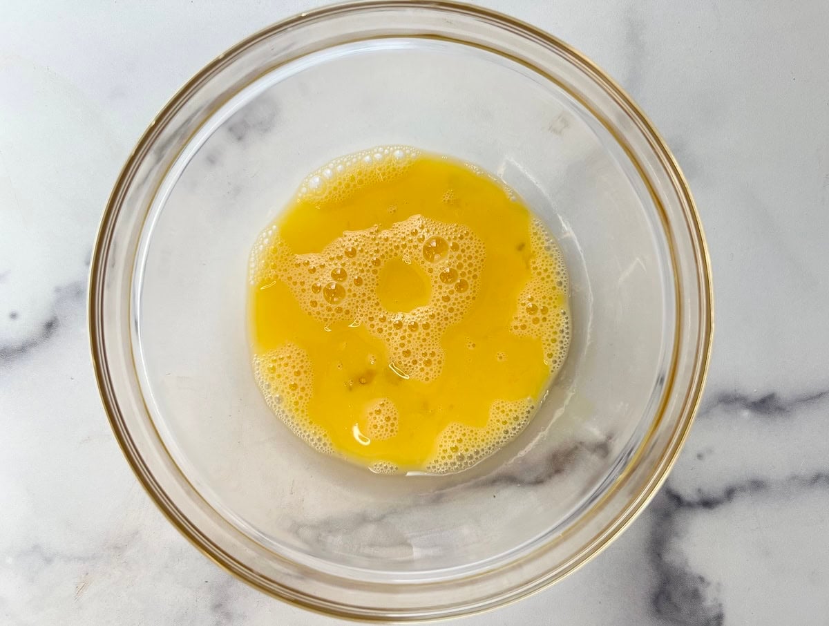 A glass bowl containing beaten eggs sits on a white marble surface.