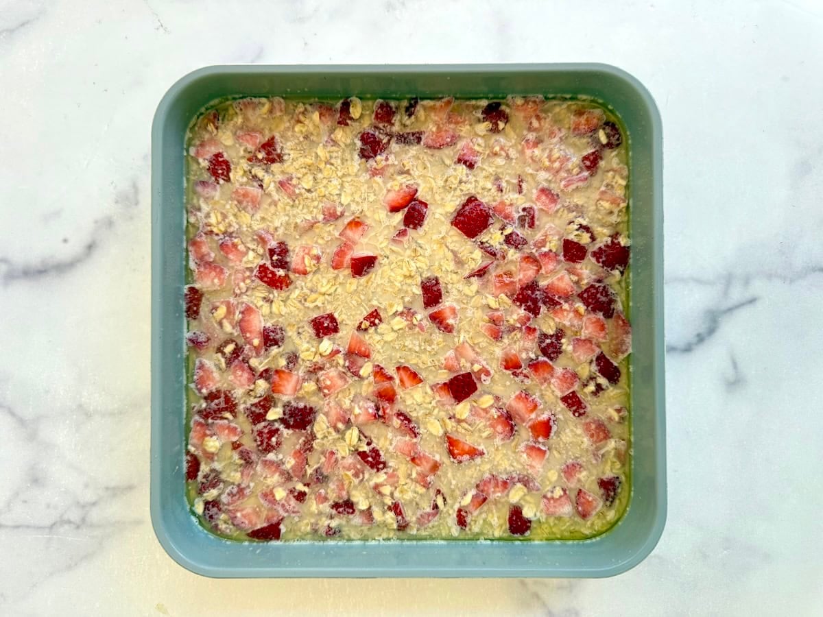 A square pan filled with an unbaked mixture of oats and chopped strawberries on a white marble surface.