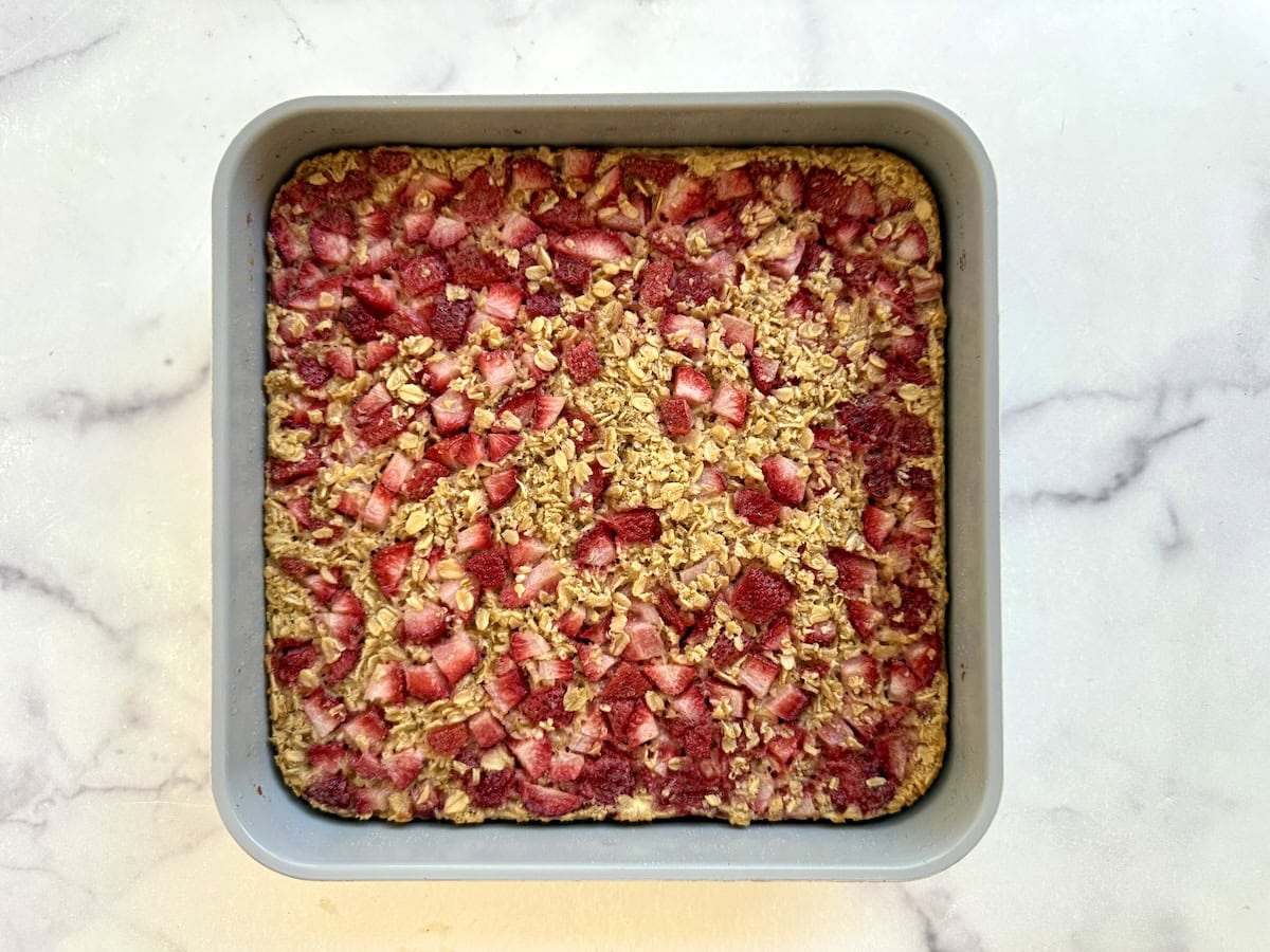 A square baking pan filled with a baked oatmeal mixture with chopped strawberries, sitting on a light marble surface.