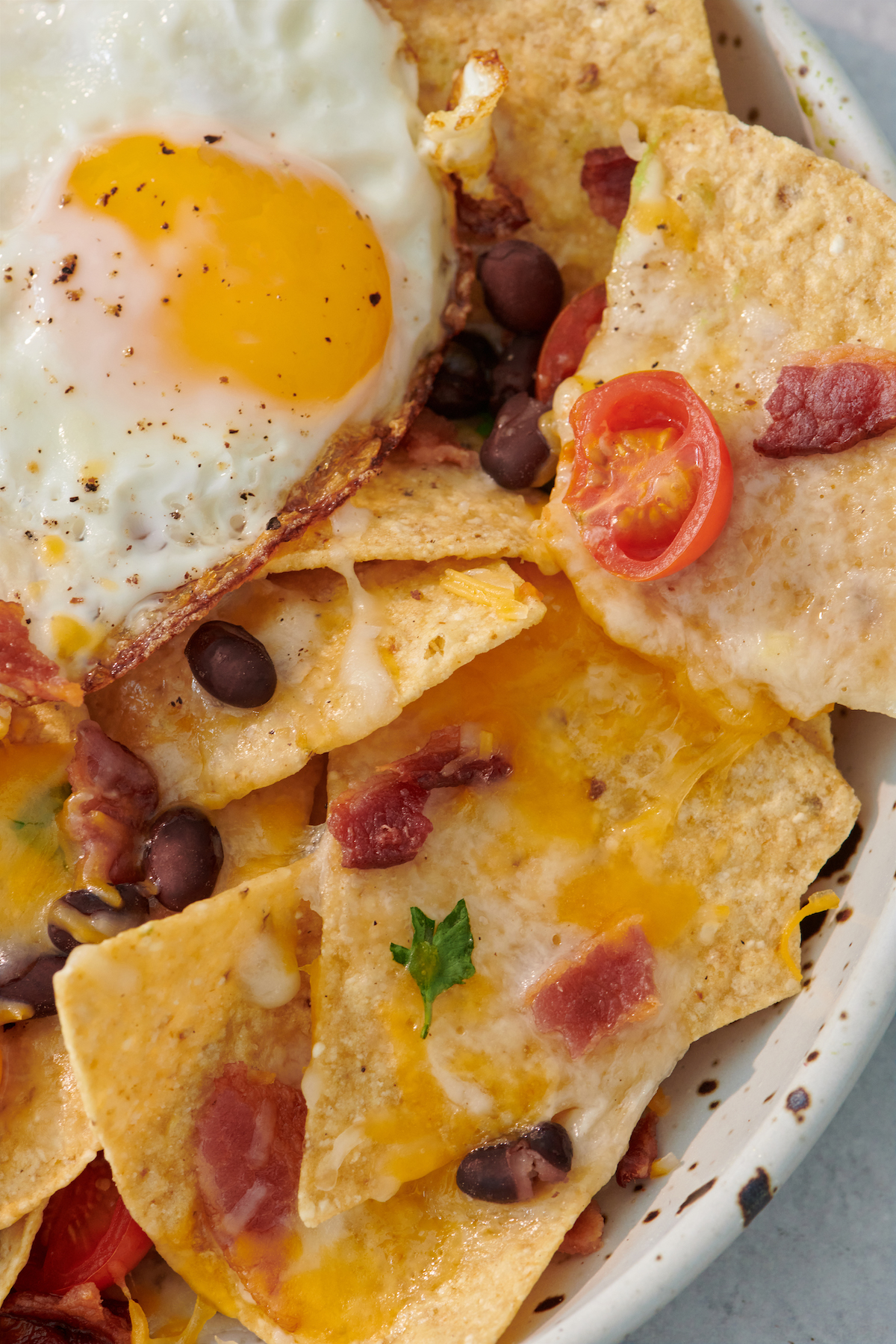 close up of breakfast nachos topped with cheese, a fried egg, beans, bacon and tomatoes