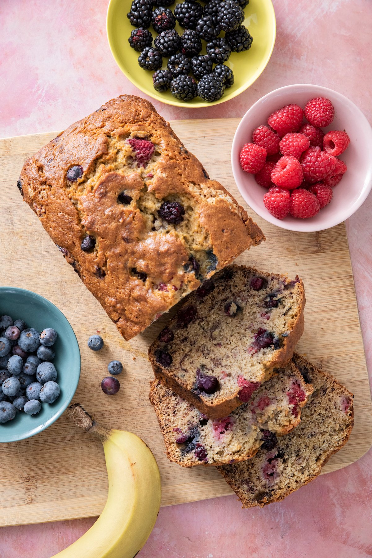 Banana Berry Bread on a serving board. The board is surrounded with three bowls with blackberries, raspberries and blueberries. 