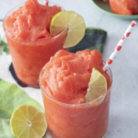 Two watermelon slushies in glasses with lime wedges.