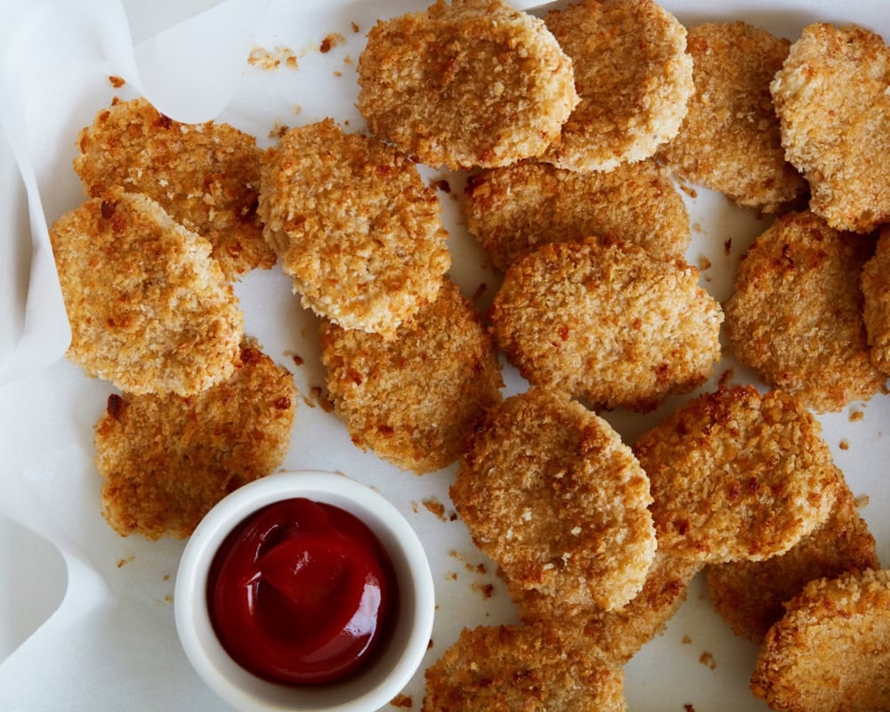 A group of breaded chicken nuggets arranged on white parchment paper next to a small cup of ketchup.