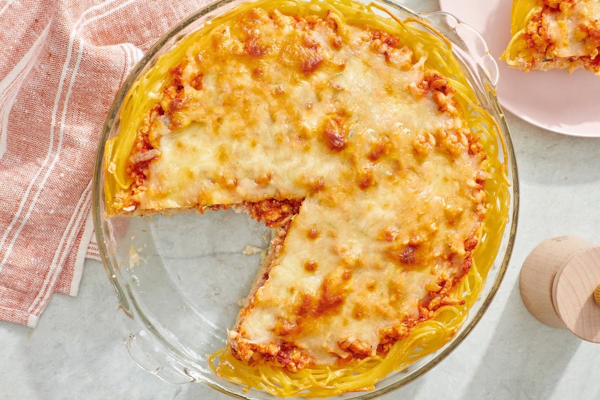 A round glass dish filled with baked spaghetti pie, topped with melted cheese and a slice missing, sits on a light surface next to a striped towel.