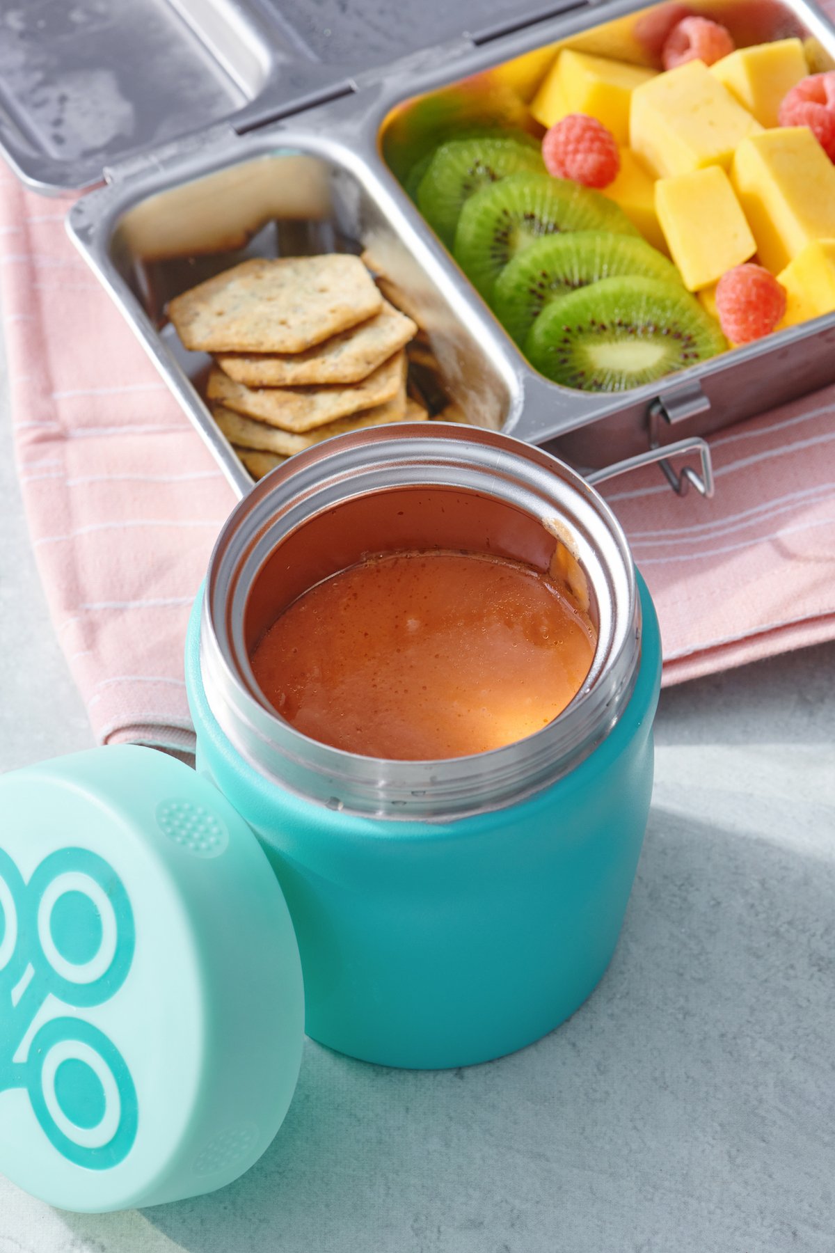 Thermos of soup in front of lunchbox with fresh fruit and crackers
