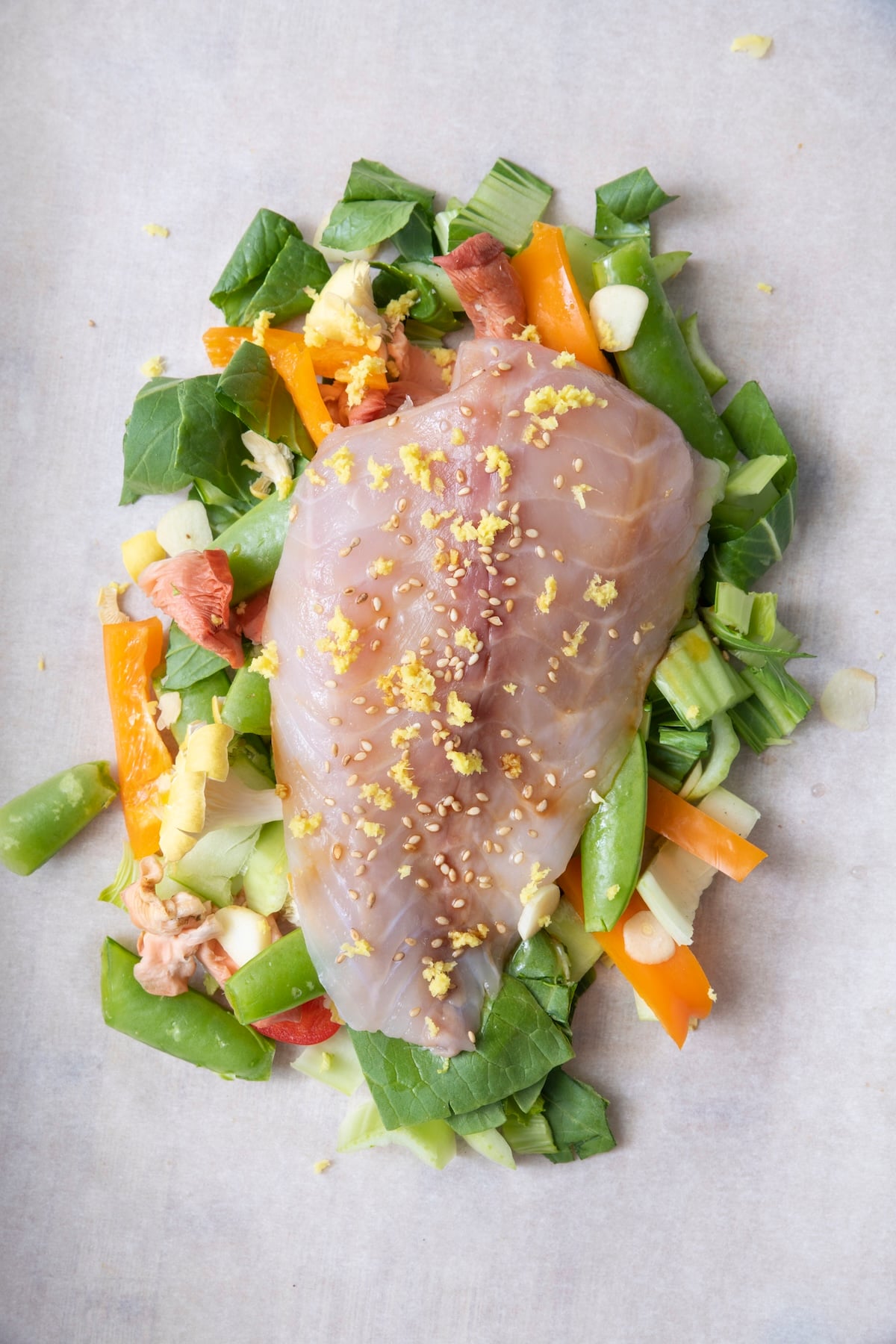 Raw whitefish fillet placed on top of chopped bok choy, snap peas, bell pepper, and shiitake mushrooms on parchment paper before sealing.