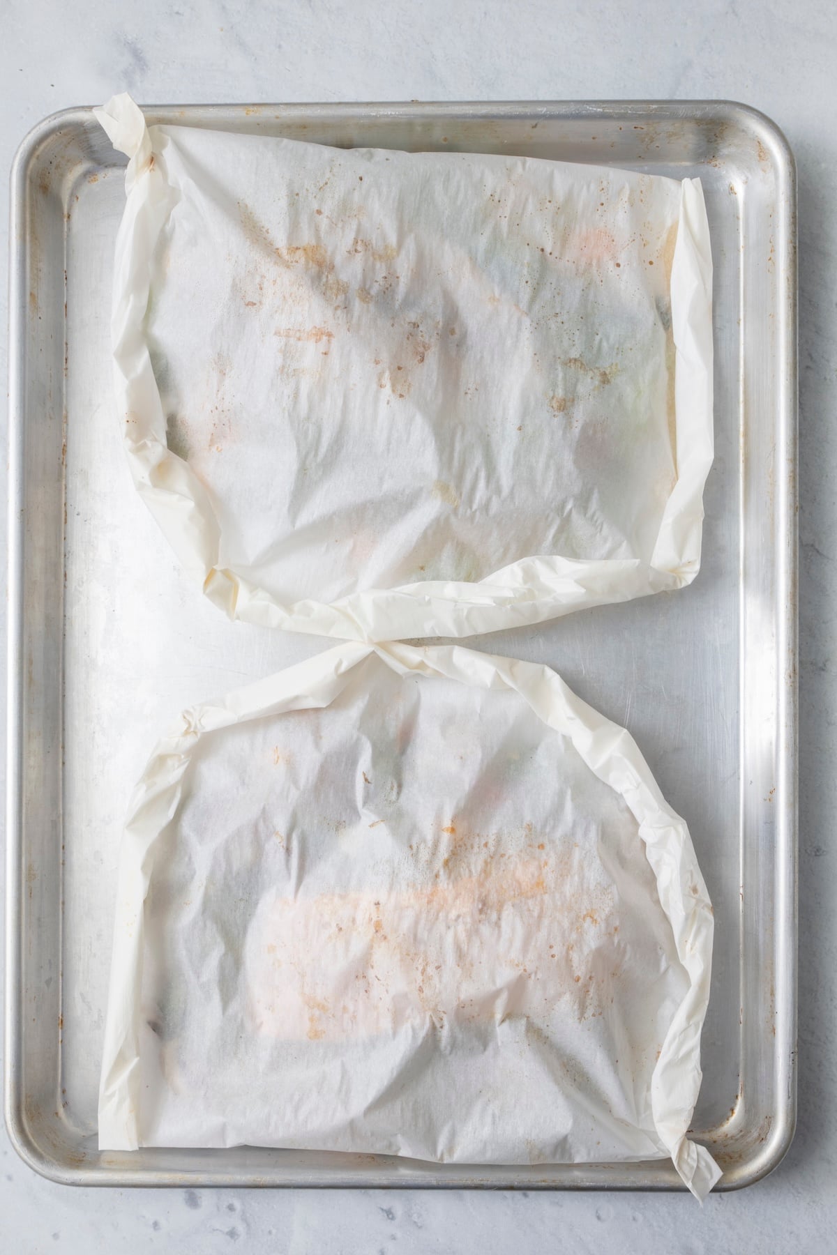 Two sealed parchment paper fish packets on an aluminum baking sheet ready for the oven.