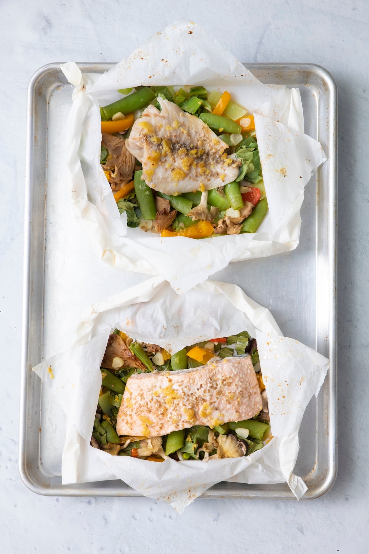 Two opened fish in parchment paper packets on a baking sheet showing cooked whitefish and salmon with vegetables after baking.
