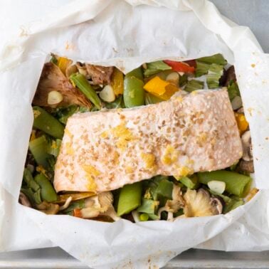 Cooked salmon fillet in an open parchment paper packet with sesame seeds, ginger, bok choy, and colorful vegetables on a baking sheet.