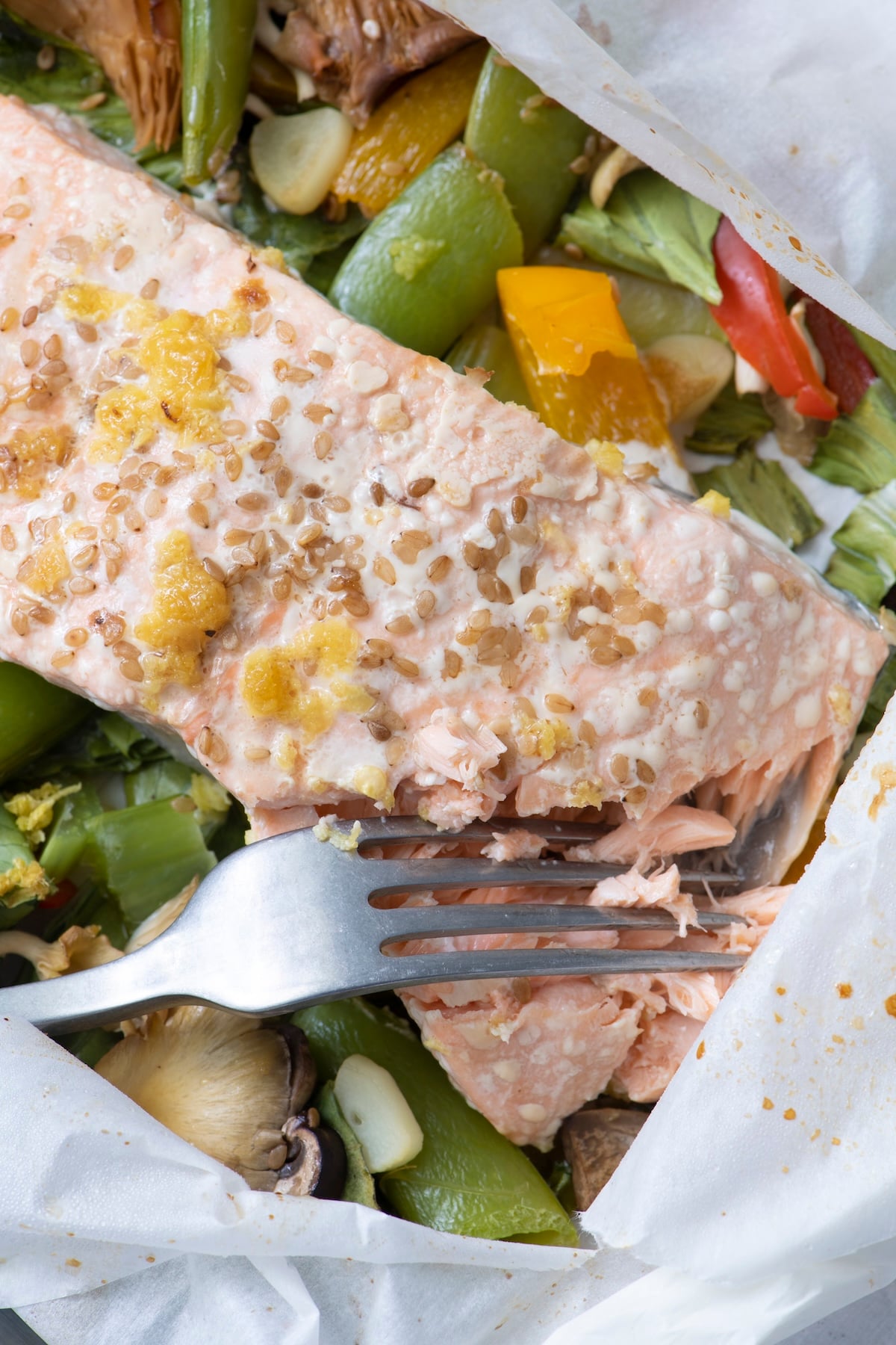 A fork flaking a perfectly cooked salmon fillet in parchment paper showing the tender flaky texture with vegetables and sesame seeds.