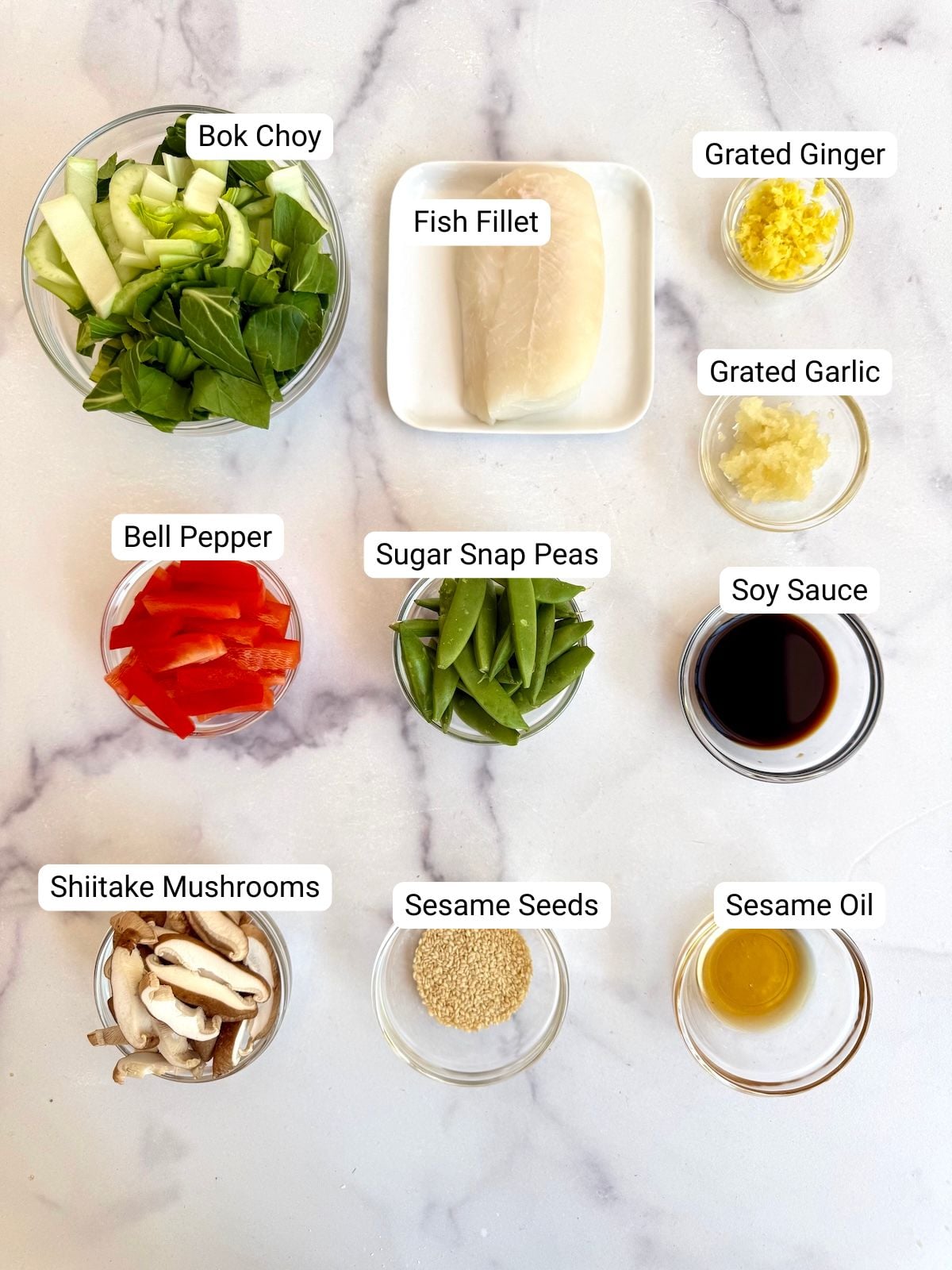 Ingredients for fish in parchment paper laid out on a marble surface including fish fillet, bok choy, bell pepper, shiitake mushrooms, sugar snap peas, grated ginger, grated garlic, soy sauce, sesame oil, and sesame seeds.
