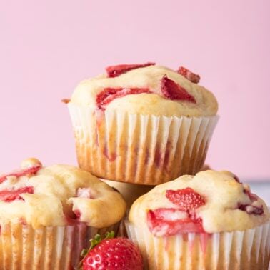 Three freshly baked strawberry muffins stacked with juicy strawberry pieces on top and a fresh strawberry in front on a white surface.