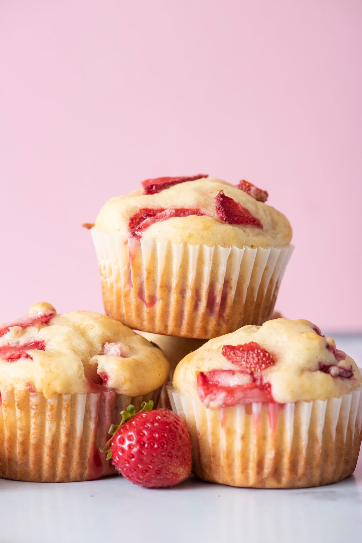 Three freshly baked strawberry muffins stacked with juicy strawberry pieces on top and a fresh strawberry in front on a white surface.