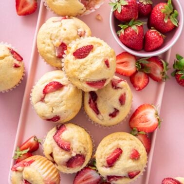 Overhead view of strawberry muffins on a pink tray surrounded by fresh strawberries and a honey dipper, one muffin with a bite taken out.