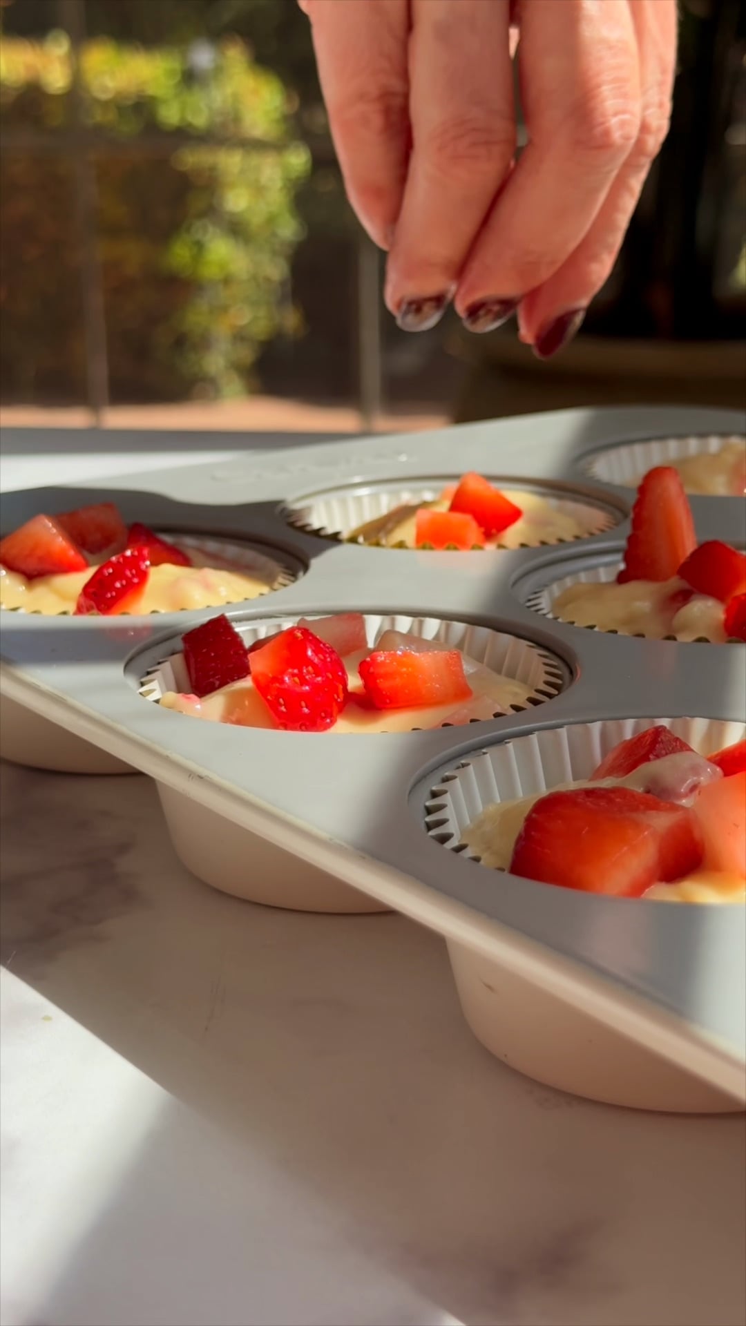 Hands pressing fresh strawberry pieces on top of filled strawberry muffin cups before baking.