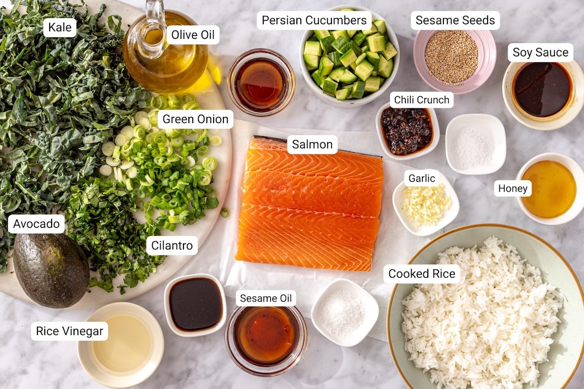 Crispy rice salmon salad ingredients including salmon fillet, jasmine rice, kale, avocado, cucumber, chili crisp, sesame oil, and soy sauce arranged on a marble surface.