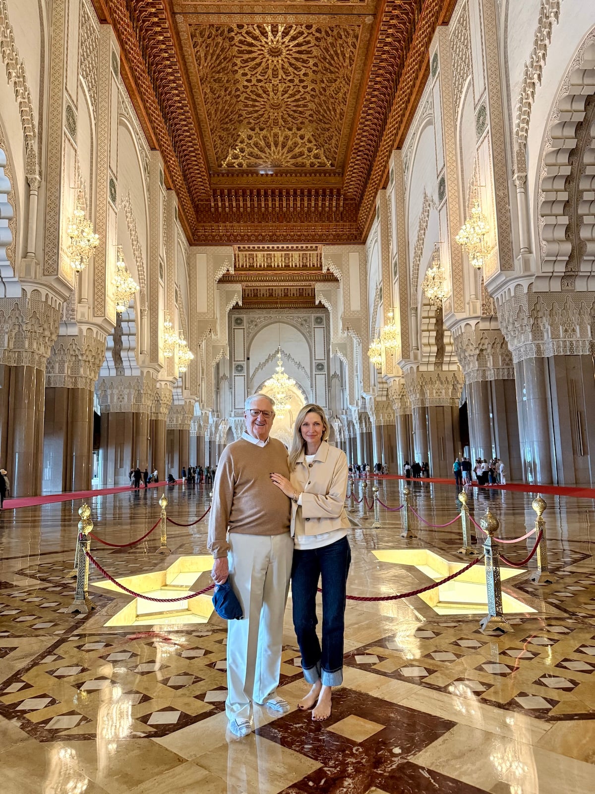 Catherine and her dad standing inside the Hassan II Mosque in Casablanca, the grand carved ceiling, marble floors, and golden chandeliers stretching behind them