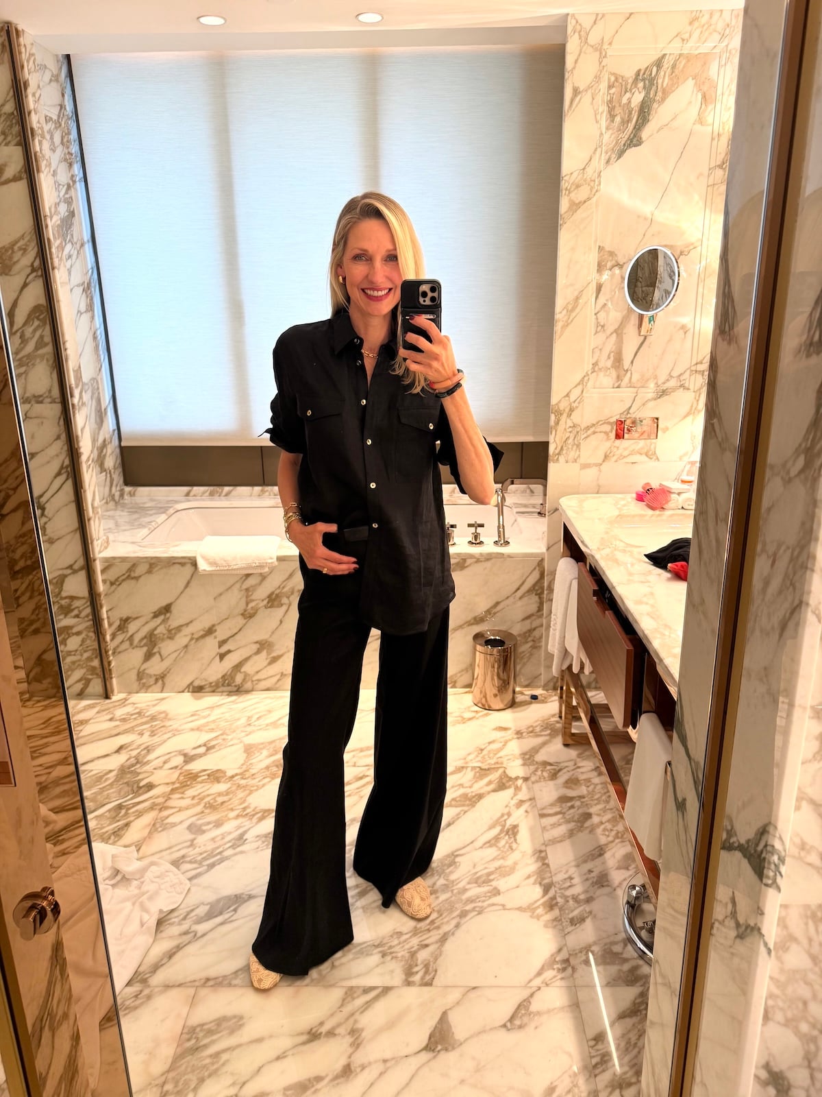 Catherine taking a mirror selfie in a black linen shirt and wide-leg trousers in a marble hotel bathroom.