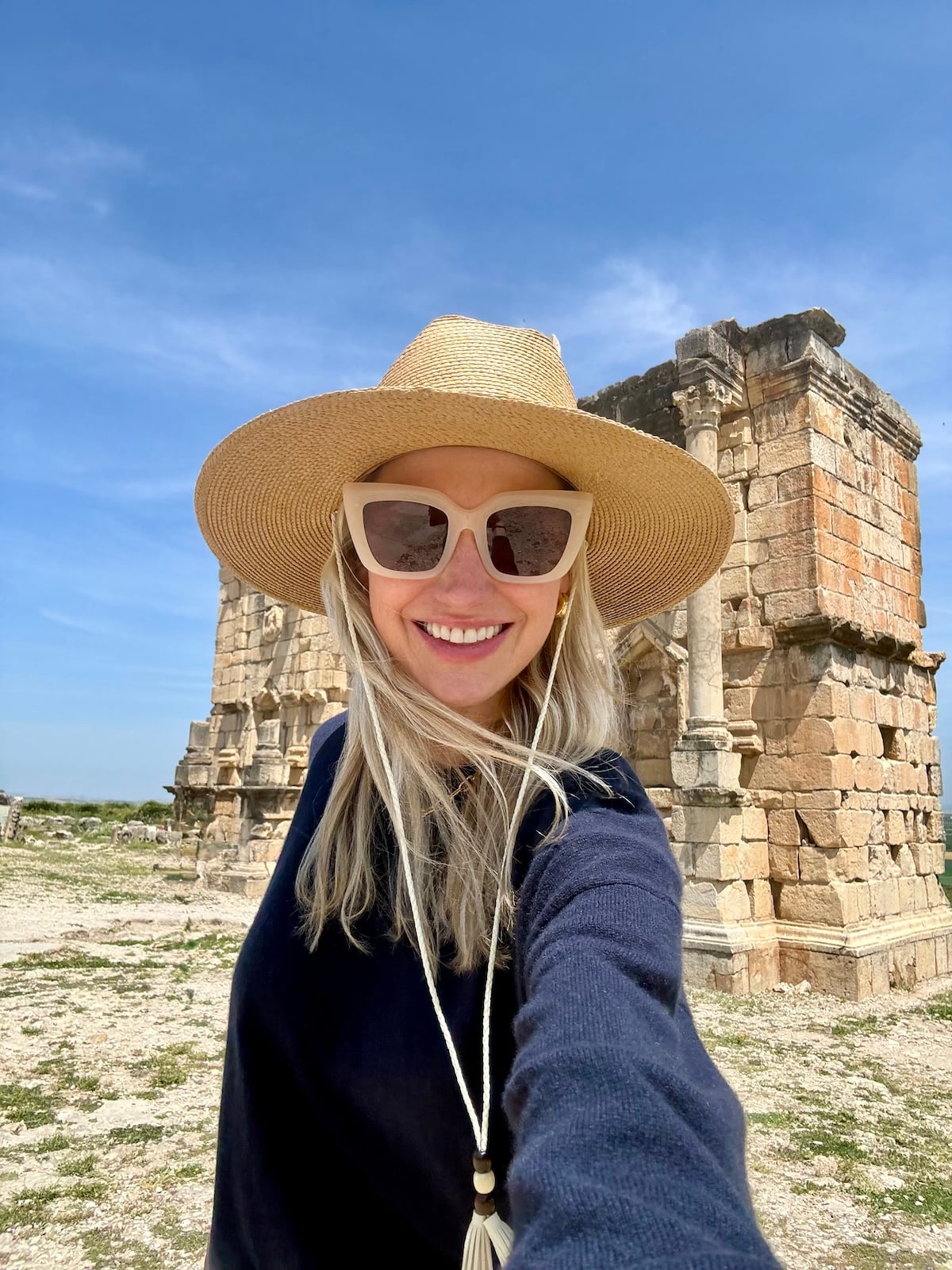 Catherine taking a selfie at the Roman ruins of Volubilis, wearing a straw hat and sunglasses, ancient stone columns behind her.