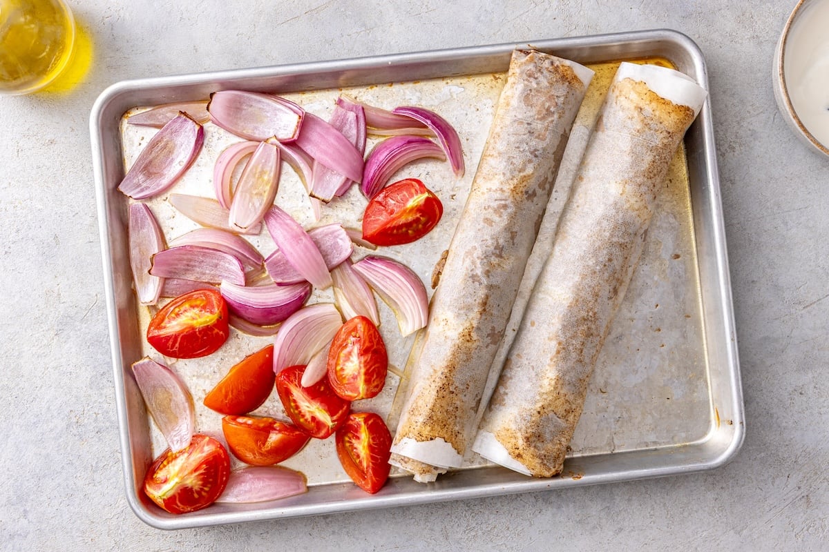 Doner kebab rolls and roasted tomatoes and red onion on a sheet pan after baking.