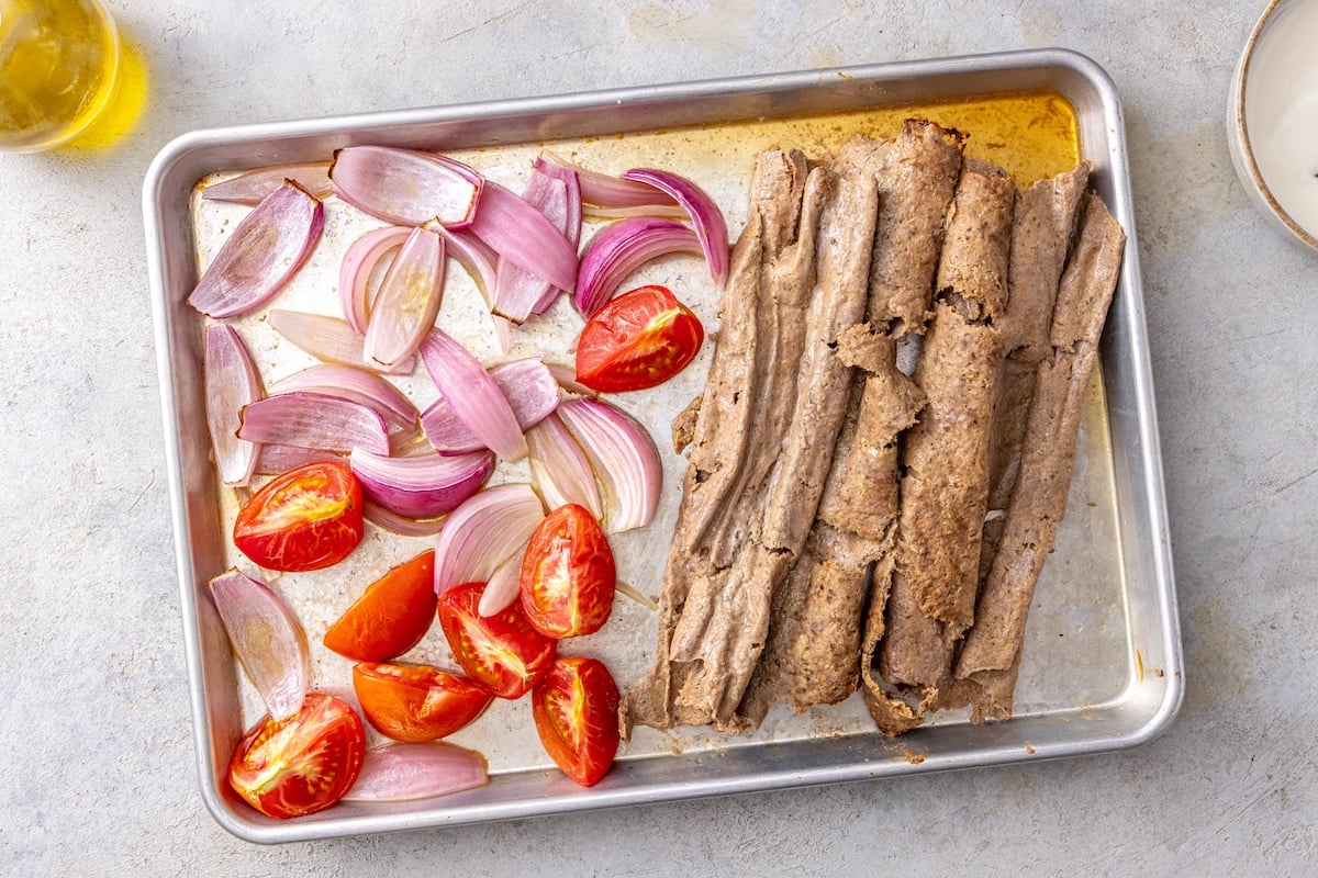 Baked doner kebab meat unrolled from parchment paper on a sheet pan with roasted tomatoes and red onion.