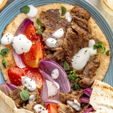 Close up of homemade doner kebab on open faced pita with crispy spiced meat, roasted tomatoes, red onion, and yogurt sauce.