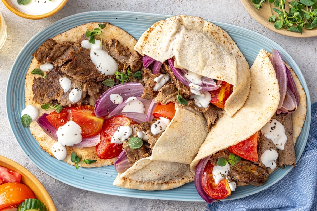 Homemade doner kebab served on warm pita bread with roasted tomatoes, red onion, and lemony yogurt sauce on a blue platter.