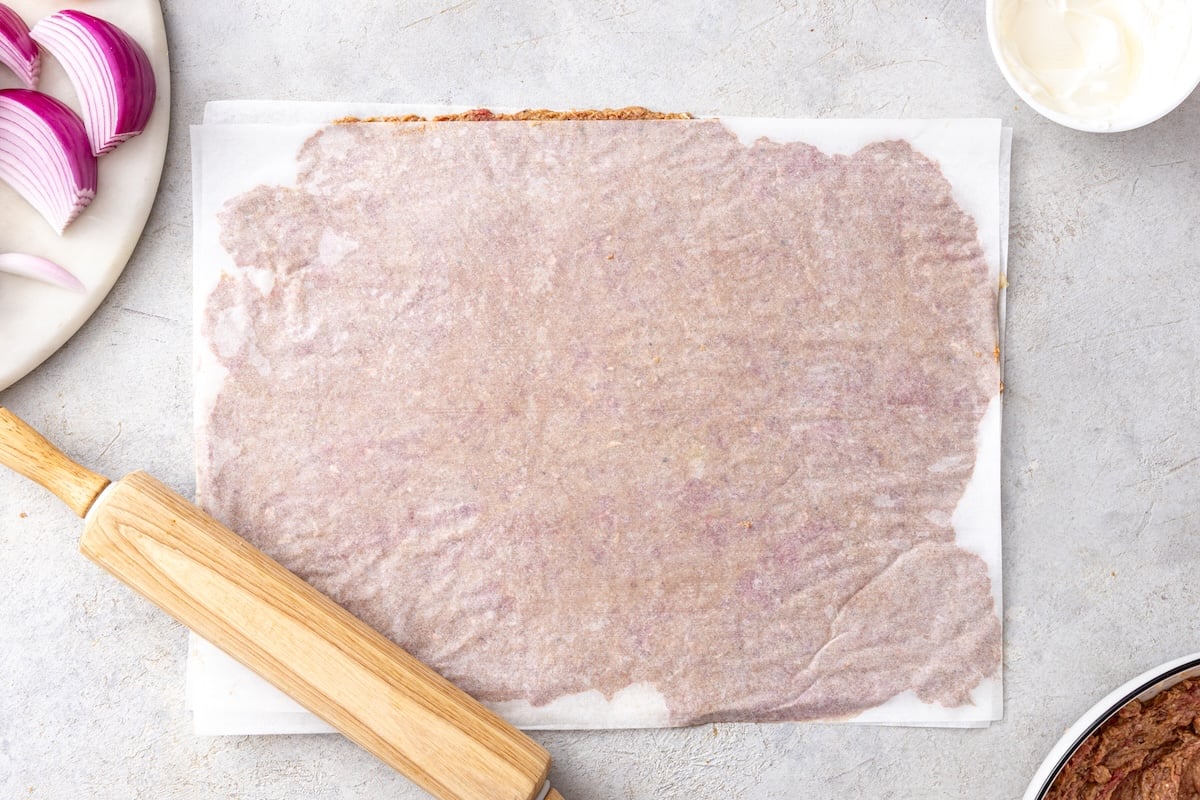 Seasoned ground beef rolled thin between two sheets of parchment paper with a wooden rolling pin for homemade doner kebab.