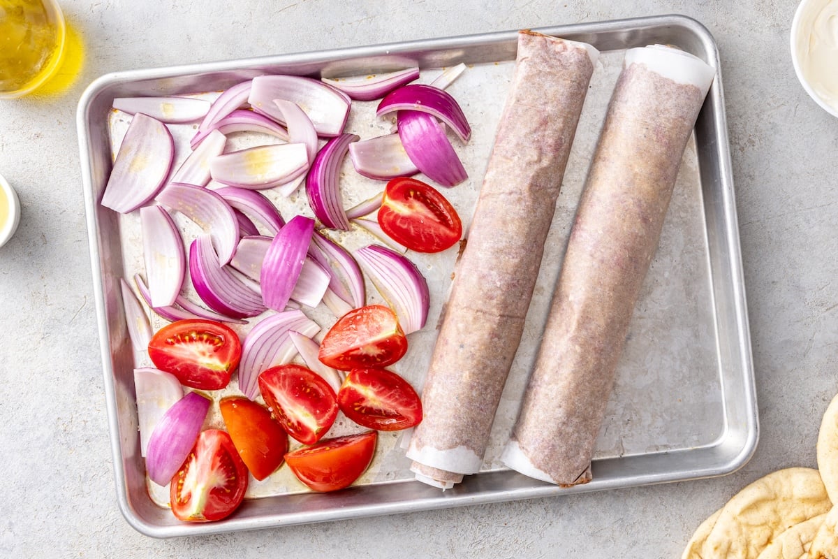 Two parchment wrapped doner kebab rolls with tomato wedges and red onion on a half sheet pan before baking.