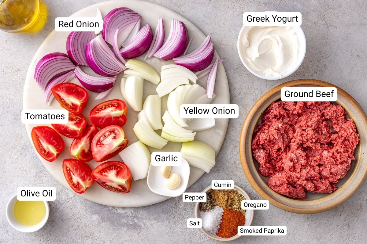 Homemade doner kebab ingredients including ground beef, yellow onion, red onion, tomatoes, garlic, Greek yogurt, olive oil, and spices arranged on a marble board.