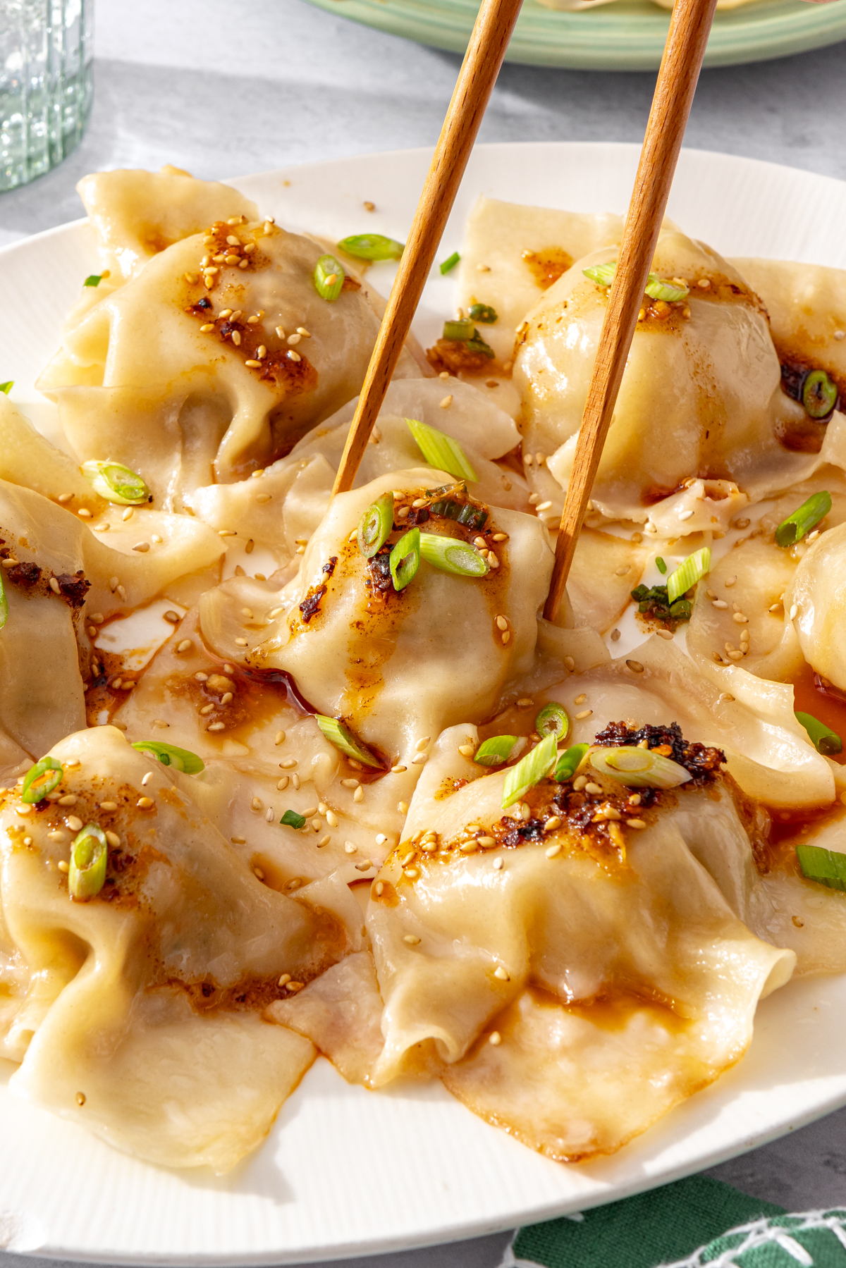 Chopsticks lifting a blanket dumpling from a plate topped with chili crisp and scallions.