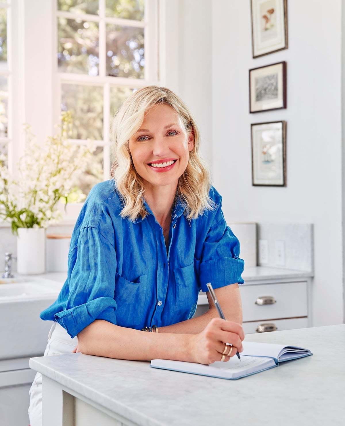 Catherine McCord in a bright blue shirt sits at a kitchen counter, writing in a notebook.