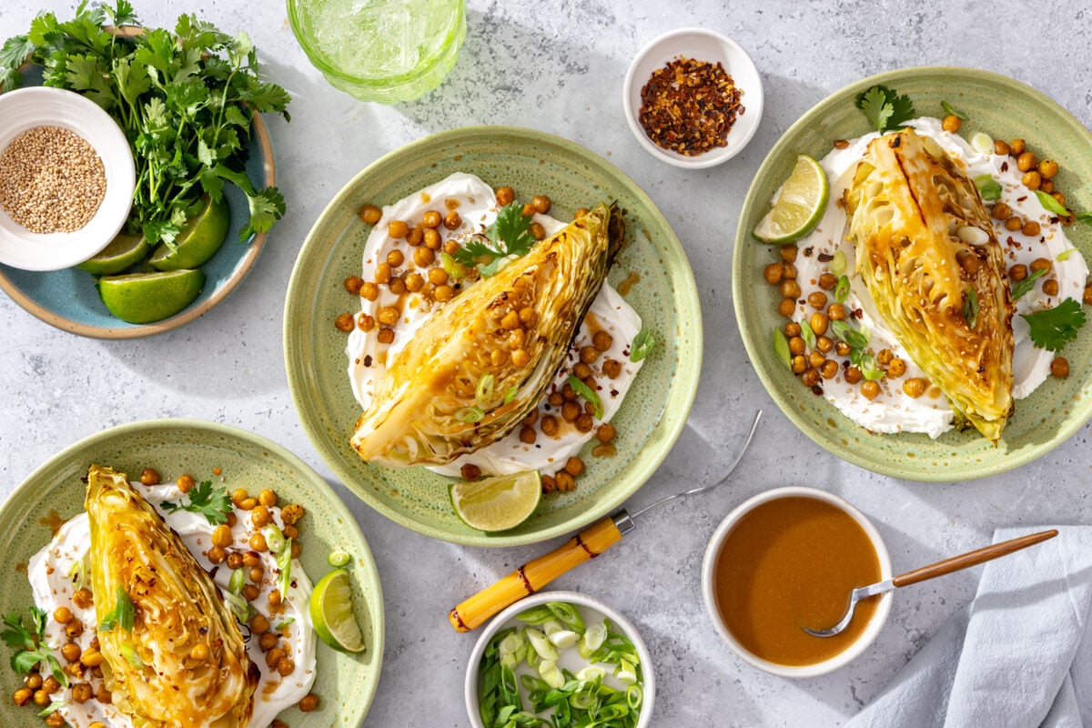 Three plates of roasted cabbage on labneh, topped with chickpeas and herbs. Surrounding bowls hold lime wedges, cilantro, sesame seeds, scallions, and a brown dressing.