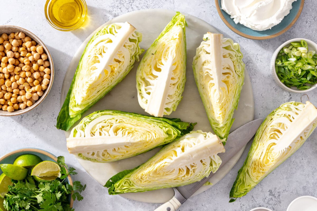 Cabbage wedges on a round cutting board with a knife, surrounded by bowls of chickpeas, labneh, green onions, cilantro, lime wedges, and a small jar of oil.